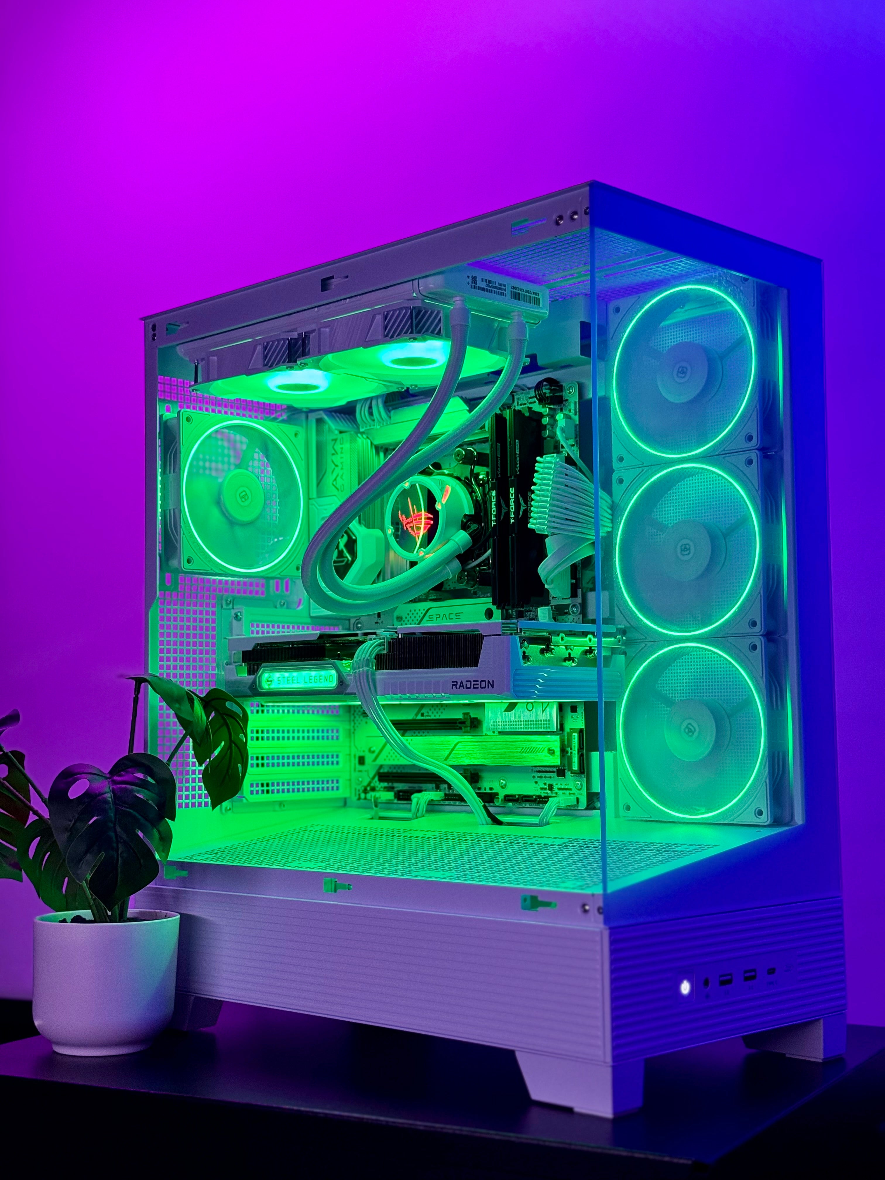 A white custom PC tower with a clear side panel, showcasing a green-lit liquid CPU cooler, graphics card, and three illuminated fans. The case is set against a purple ambient light background with a small potted plant on the left.