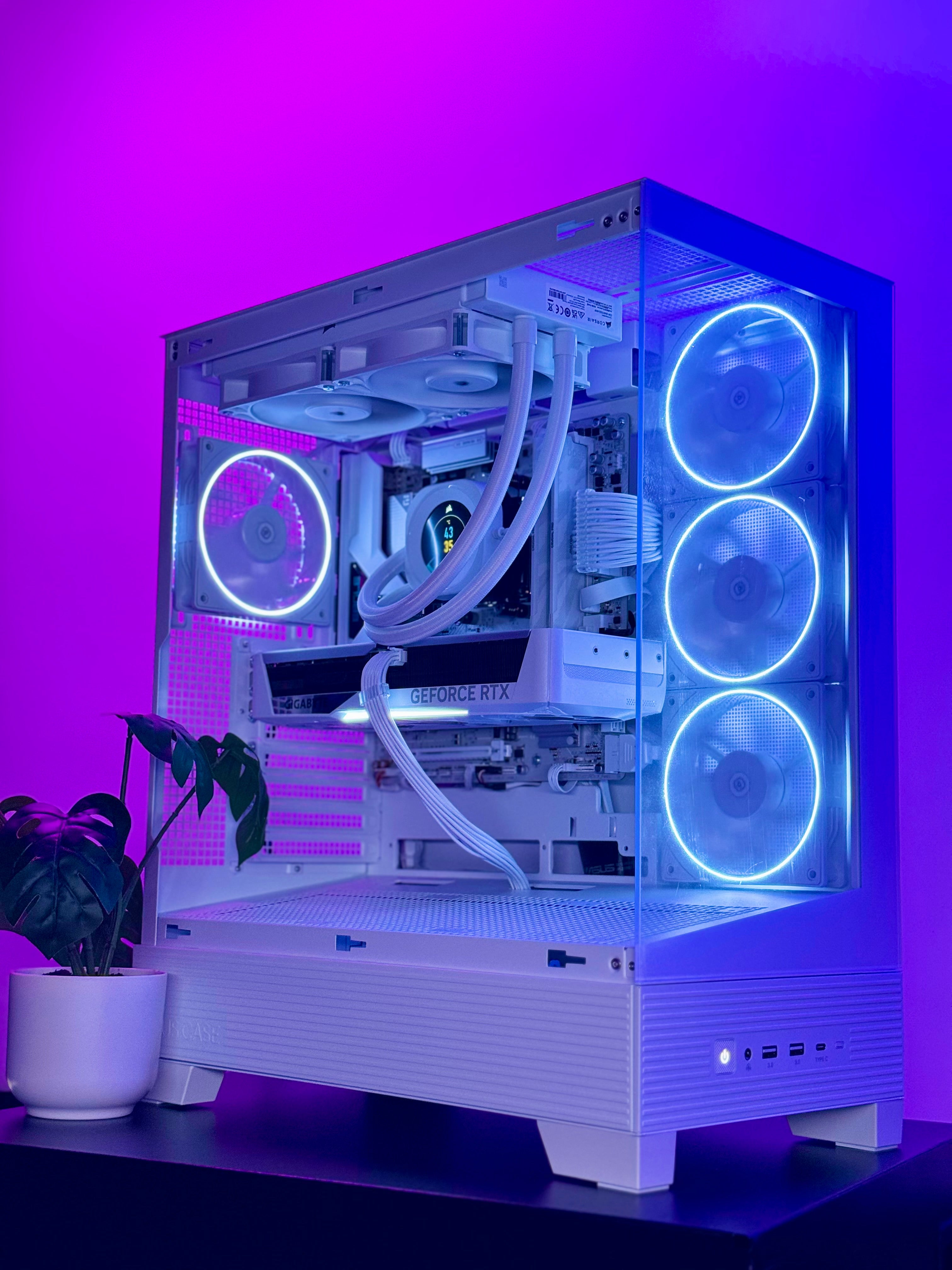 A white custom PC tower with a transparent side panel, illuminated by glowing blue LED fans and RGB lighting, set against a purple ambient background with a small potted plant on the left.
