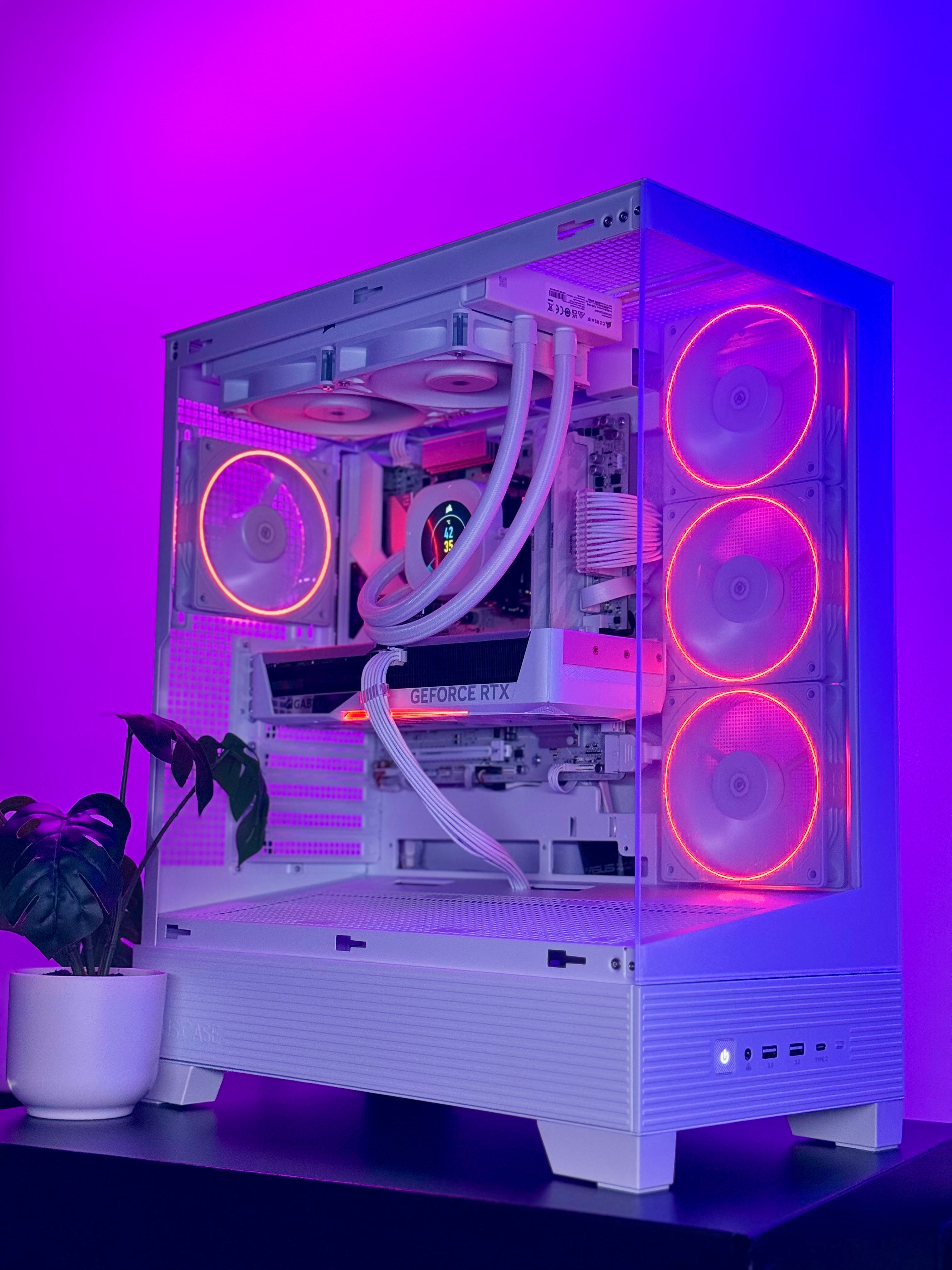 A white custom-built desktop PC tower with a transparent side panel, showcasing a white liquid CPU cooler, white cables, and multiple RGB fans glowing with pink and orange light. The PC is set on a black surface against a purple LED-lit background, with a potted plant on the left.