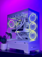 A white custom-built desktop PC tower with a clear side panel, showcasing a white liquid CPU cooler, white fans, and a visible NVIDIA RTX graphics card. The PC is illuminated by yellow and green RGB lighting against a purple background, sitting on a black surface next to a potted plant.