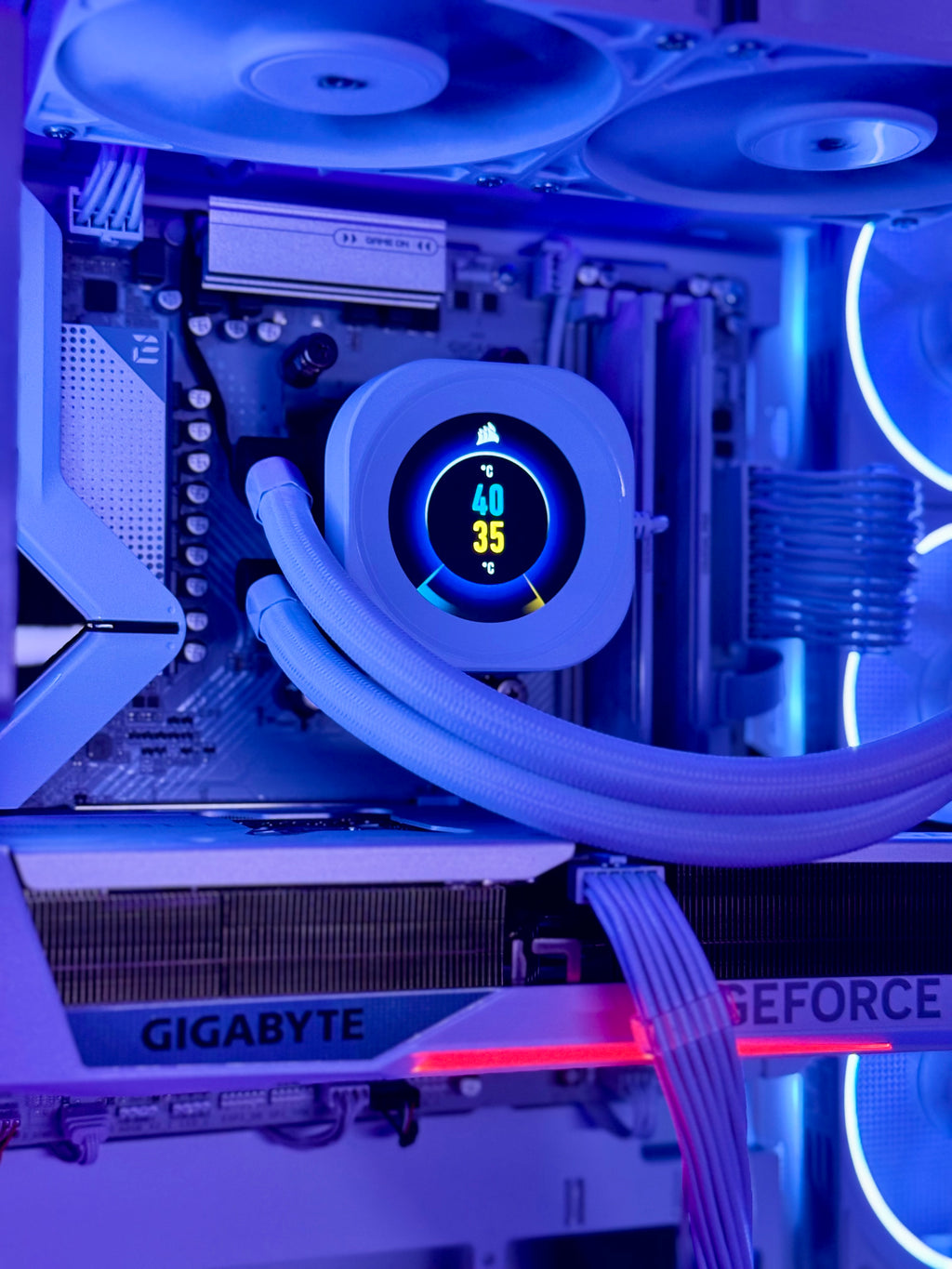 A close-up of a custom PC build with a liquid CPU cooler featuring a circular digital temperature display, illuminated by blue LED lighting. Visible components include a Gigabyte Z790 AORUS motherboard and a GeForce RTX graphics card.