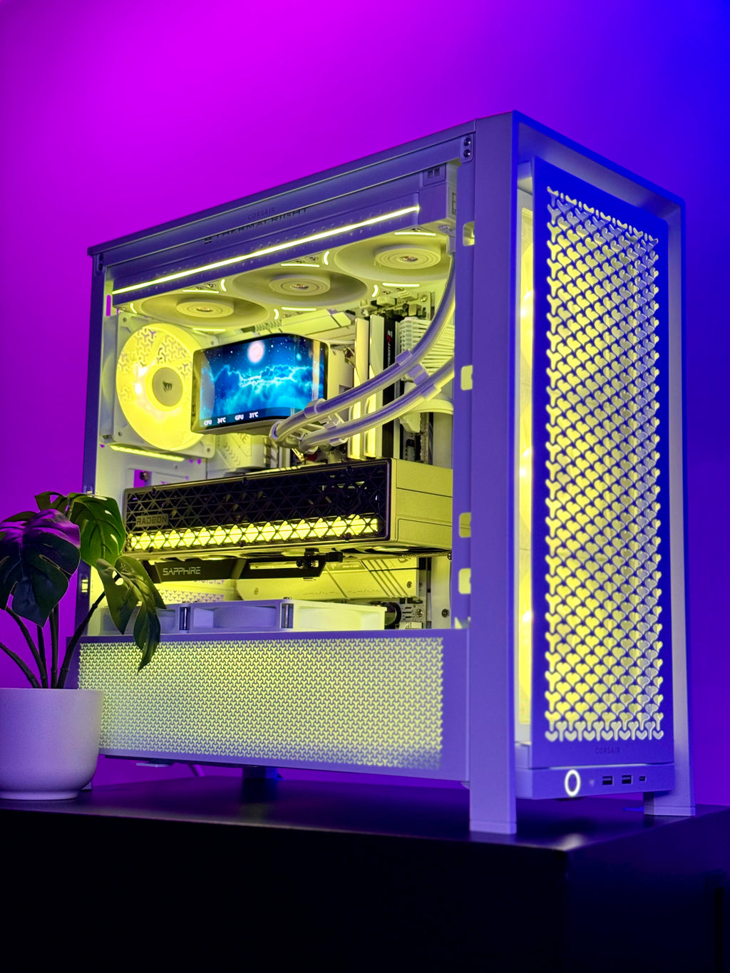 A custom white PC case with a glass side panel, illuminated by yellow and blue LED lighting, showcasing a liquid CPU cooler and components inside. The case is set on a dark surface with a potted plant on the left, against a purple-lit background.