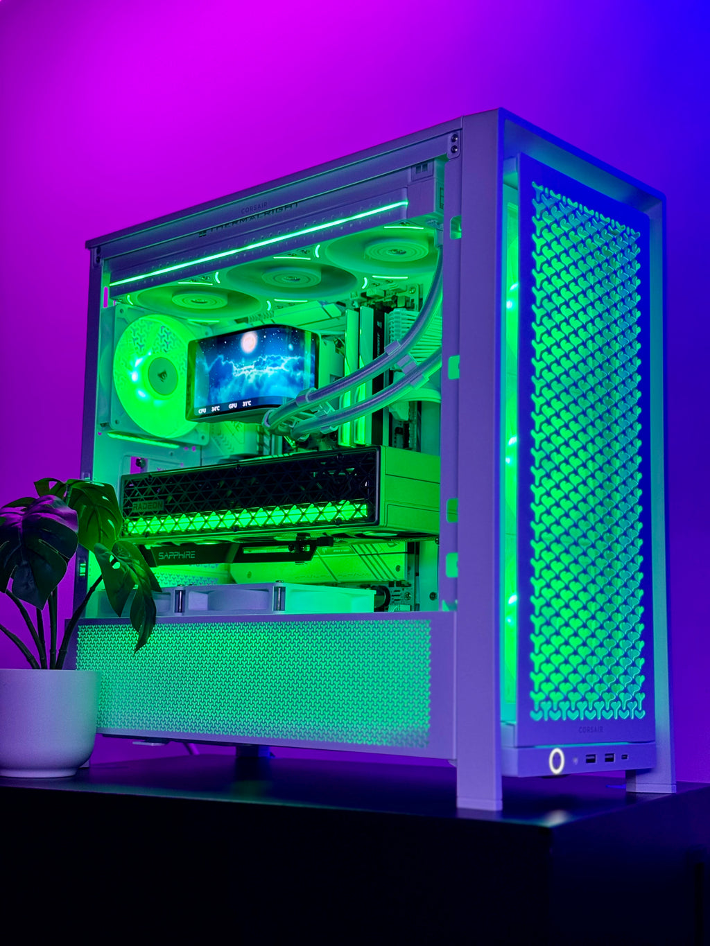 A custom white desktop PC tower with a clear side panel, showcasing a green-lit internal liquid CPU cooler with fans and tubing. The PC sits on a dark surface against a purple and pink ambient light background, with a potted plant on the left.