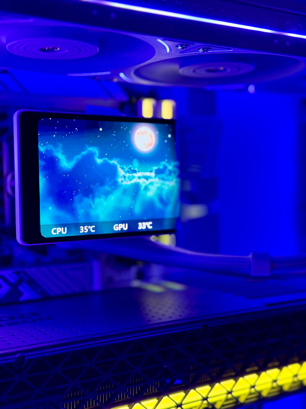 A close-up of a computer case interior illuminated by blue LED lighting, showing a front-mounted display screen with a moon wallpaper and temperature readings for CPU and GPU, along with yellow light strips along the bottom.