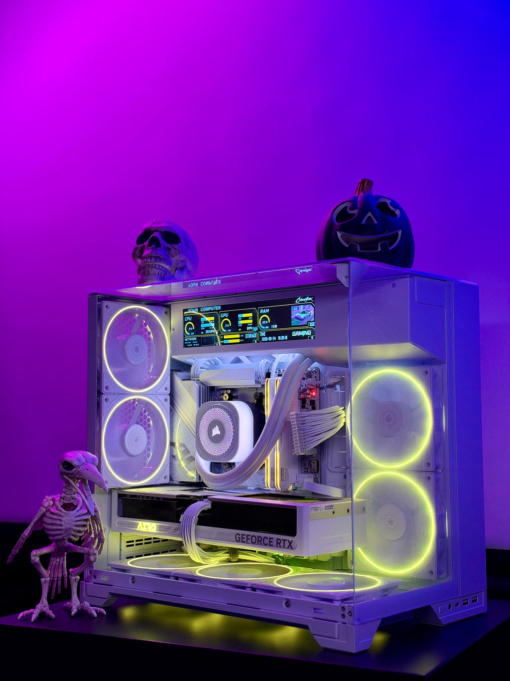 A custom white PC tower with glowing yellow and white RGB fans, illuminated by purple and blue LED lighting against a purple background. The PC is decorated with a skull, a jack-o'-lantern, and a small skeleton figurine.