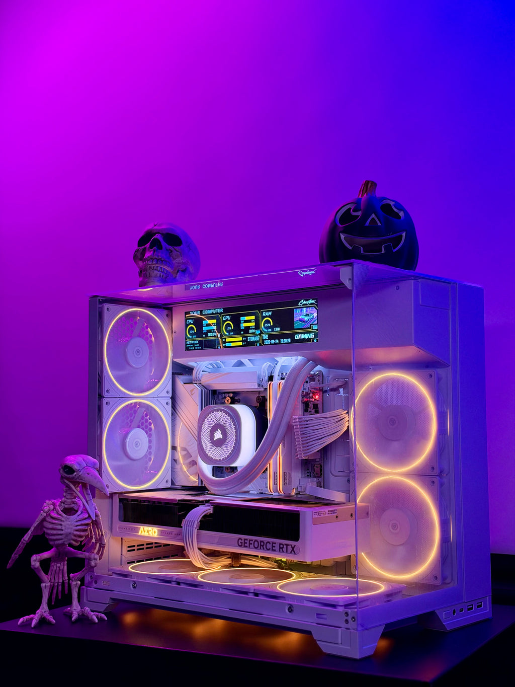 A custom white PC case with a transparent side panel, illuminated by glowing orange and white fans and internal LED lighting. The case is decorated with a small skeleton figurine on the left, a skull on top, and a jack-o'-lantern on top, set against a purple and blue gradient background.