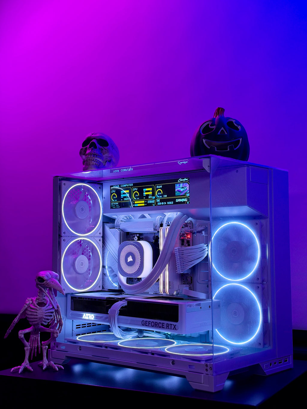 A custom white PC tower with a glass side panel, illuminated by blue and purple LED fans and internal lighting. The PC is styled with Halloween decorations, including a skull, a jack-o'-lantern, and a small skeleton figurine, set against a vibrant purple and blue gradient background.