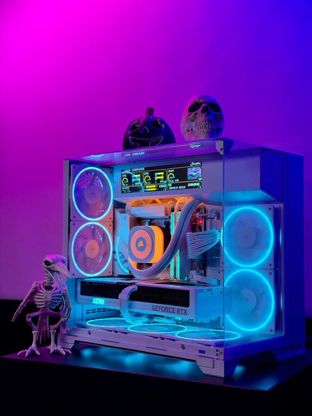 A custom PC tower with a transparent side panel, showcasing a liquid CPU cooler, GeForce RTX graphics card, and multiple RGB fans glowing in blue and orange. The PC sits on a black surface against a vibrant pink and purple gradient background, decorated with Halloween props including a skeleton figurine, a jack-o'-lantern, and a skull.