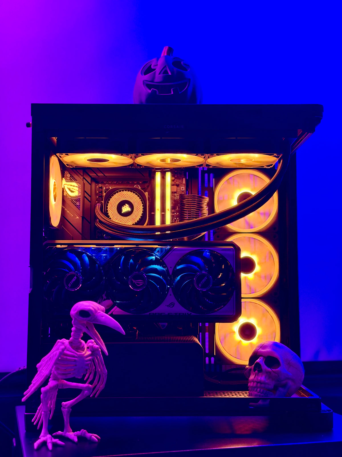 A custom PC tower with a transparent side panel, illuminated by glowing orange and yellow fans and internal LED lights, styled with a skeleton raven, a skull, and a jack-o'-lantern on a blue and purple background.