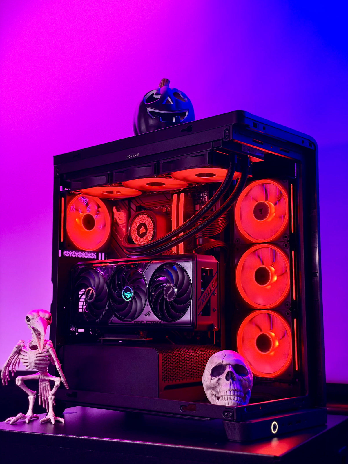 A black PC case with a glass side panel, illuminated by glowing red and orange fans and RGB lighting. The case is styled with Halloween decorations, including a skeleton figurine, a skull, and a jack-o'-lantern, set against a vibrant purple and blue gradient background.