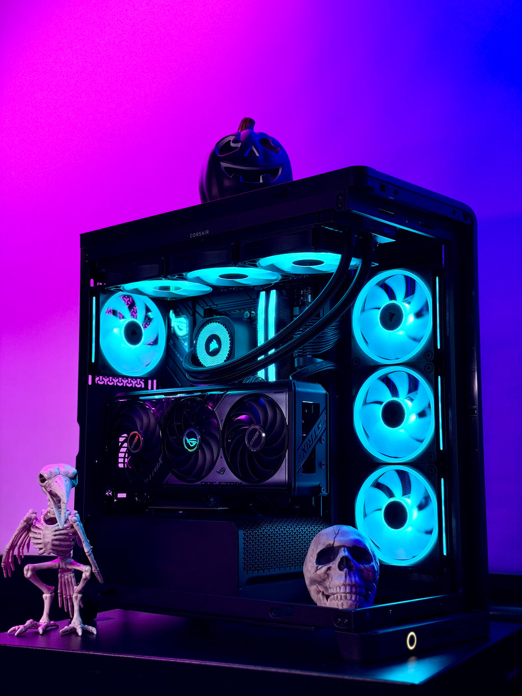 A black desktop PC case with a glass side panel, illuminated by bright blue and cyan RGB fans and internal lighting. The case is styled with Halloween decorations, including a skeleton figurine, a skull, and a jack-o'-lantern, set against a vibrant purple and pink gradient background.