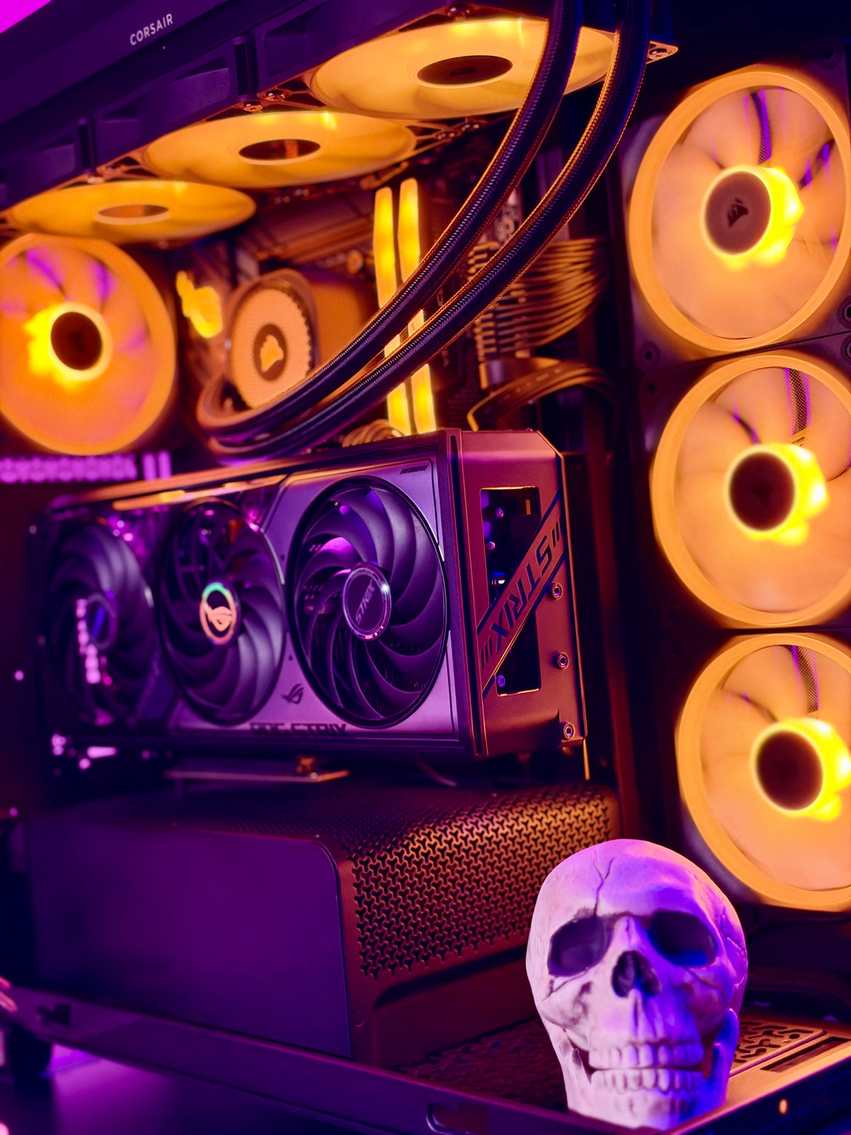 A high-performance custom PC build featuring a Corsair liquid cooler, an AMD Ryzen processor, a GeForce RTX 3080 graphics card, and multiple RGB-lit fans glowing in yellow and orange, with a small skull prop in the foreground.