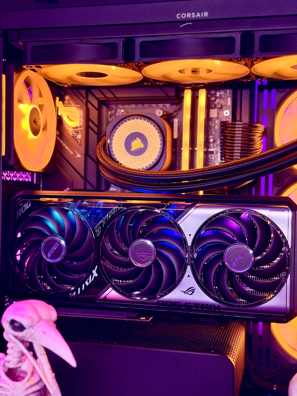 Corsair and ASRock desktop PC interior with RGB-lit fans, a large triple-fan graphics card in the foreground, and a small skull prop at the lower left.
