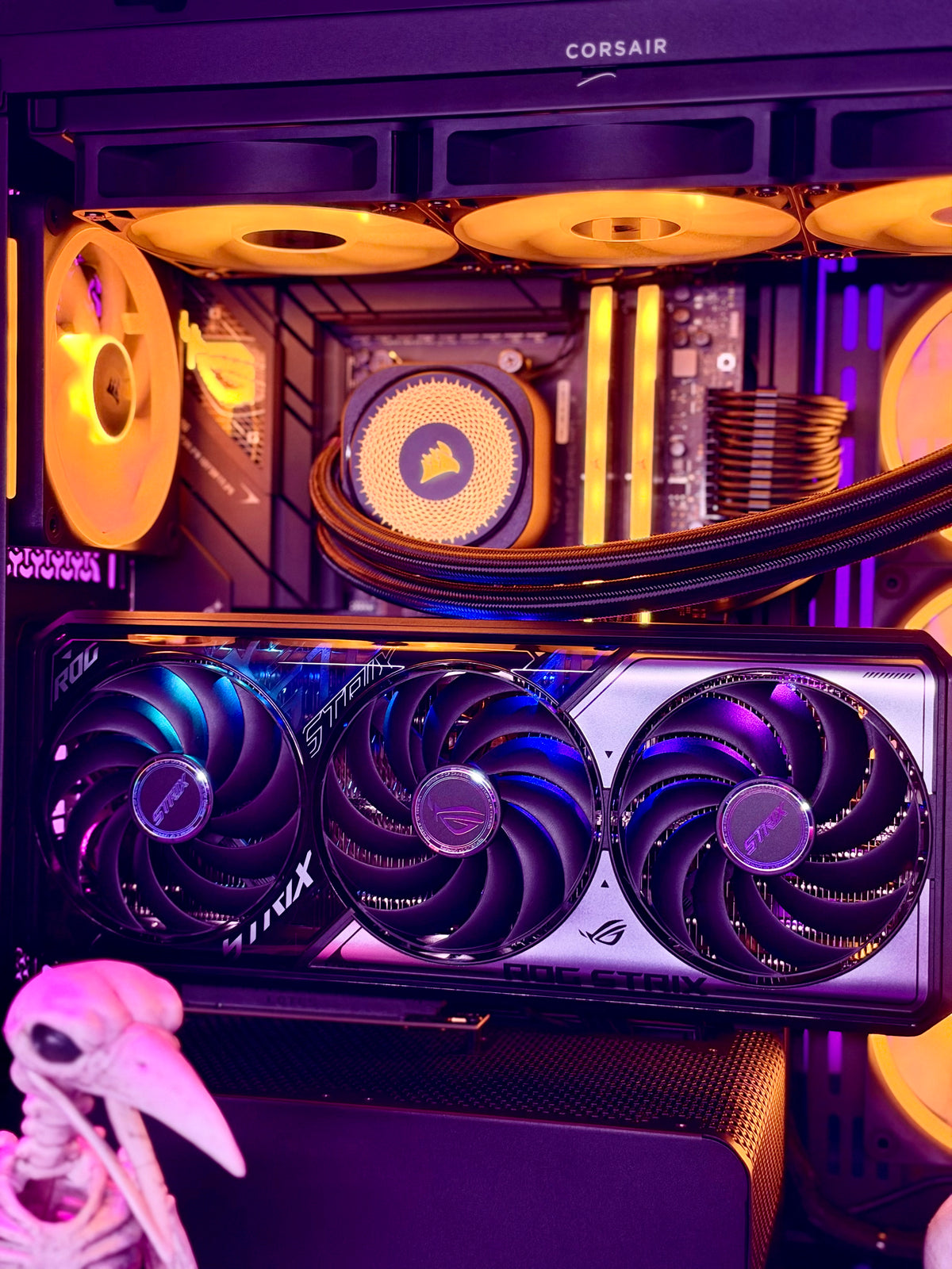 Corsair and ASRock desktop PC interior with RGB-lit fans, a large triple-fan graphics card in the foreground, and a small skull prop at the lower left.